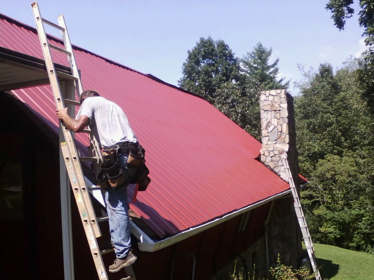 Roofer on a ladder beside a red metal roof and stone chimney — Soffit & Fascia Repair by Roofing Brothers in Dassel
